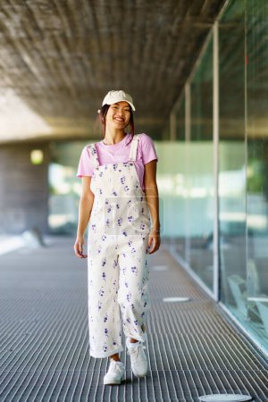 Photo for Full body happy young Asian female in stylish clothes and cap, smiling and looking at camera while walking near glass wall of contemporary building in daytime - Royalty Free Image