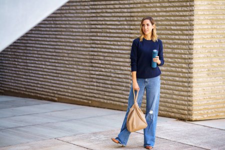 Foto de Mujer de cuerpo completo en jeans elegantes y suéter con bolsa y termo mirando hacia otro lado mientras está de pie en el pavimento fuera del edificio moderno - Imagen libre de derechos
