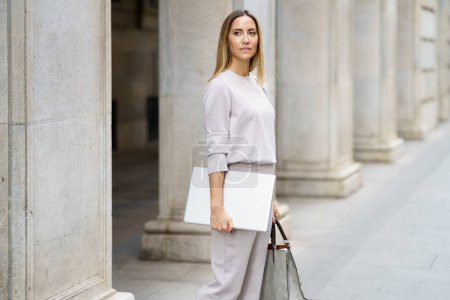 Photo for Serious self assured adult woman with blond hair in gray outfit standing on street near old building and holding laptop and handbag while looking away - Royalty Free Image