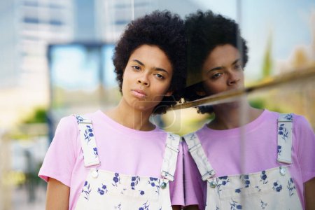 Foto de Grave joven afroamericana femenina con peinado afro mirando a la cámara mientras está de pie cerca de la casa de cristal en la calle contra el fondo borroso - Imagen libre de derechos