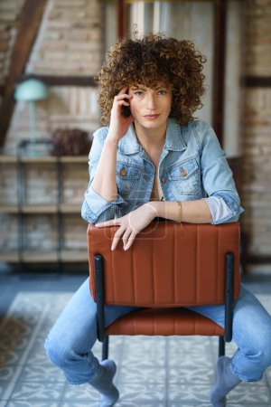 Photo for Positive young female with curly hair in jeans jacket sitting on leather chair and touching head in room with brick wall and tiled floor - Royalty Free Image