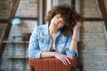 Téléchargez les photos : Jeune femme sérieuse en sous-vêtements et veste en jean assise sur une chaise en cuir et touchant les cheveux tout en regardant la caméra contre un mur de briques floues - en image libre de droit