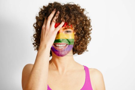 Photo for Happy curly haired female with painted in LGBT colors face covering eye looking at camera against white background equality and tolerance concept - Royalty Free Image