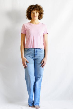 Téléchargez les photos : Corps complet de femme aux cheveux bouclés en jeans et t-shirt rose appuyé sur un mur blanc regardant la caméra - en image libre de droit