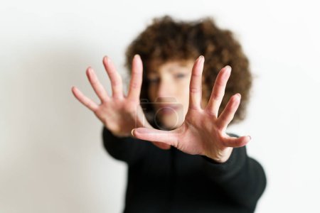 Photo for Defocused face of girl model, with curly hair looking at camera and showing stop gesture with full open palms and fingers against gray background in blurring unseen side illuminating lights - Royalty Free Image