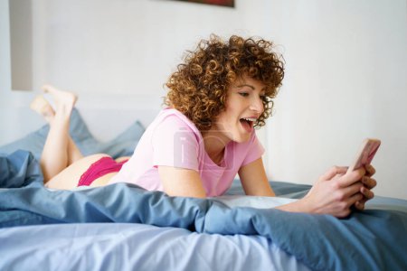 Téléchargez les photos : Vue latérale complète du corps de la jeune femme joyeuse avec les cheveux foncés bouclés couchés sur le lit et la navigation téléphone mobile pendant le week-end à la maison - en image libre de droit