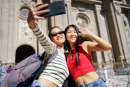 Photo for Cheerful young Asian female tourists in casual outfits smiling and taking self portrait on smartphone while standing against Cathedral of Granada in Spain during holidays - Royalty Free Image