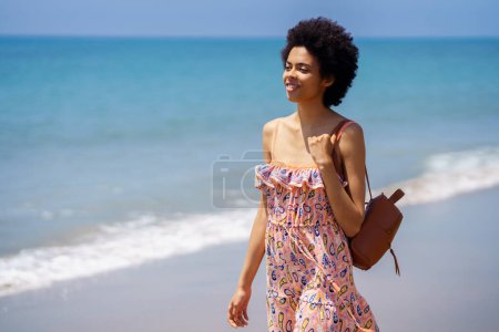 Photo for Black woman in colorful dress smiling while exploring sandy beach near rippling ocean and looking away - Royalty Free Image