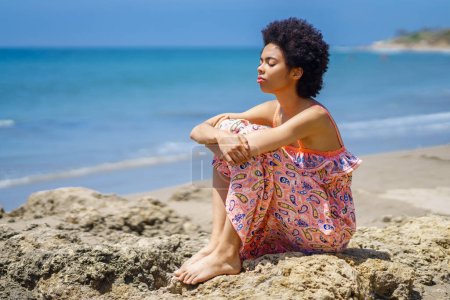 Photo for Barefoot black woman in ornamental dress with curly hair, embracing knees and inhaling fresh air with closed eyes while chilling on rough rocks against waving sea on resort - Royalty Free Image