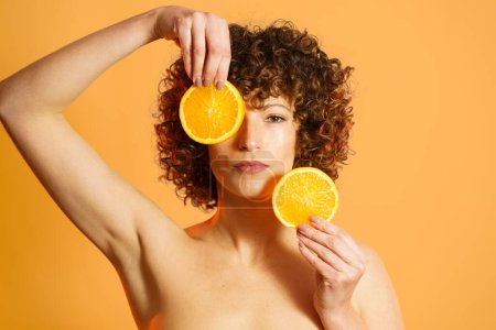 Photo for Serious adult female with curly hair covering face with slices of ripe citrus during beauty routine against orange background - Royalty Free Image