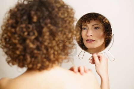 Foto de Hermosa mujer joven, con el pelo rizado y maquillaje de pie con la espalda contra la cámara mientras sostiene el espejo en la mano y mirando hacia otro lado en reflejo de sí mismo cerca de fondo gris - Imagen libre de derechos