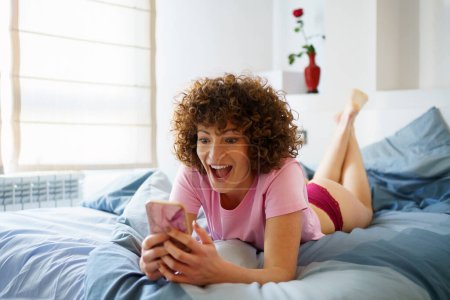 Téléchargez les photos : Délicieuse jeune femme aux cheveux bouclés couchée sur un lit confortable tout en surfant sur les médias sociaux via téléphone portable dans la chambre à coucher à la maison - en image libre de droit