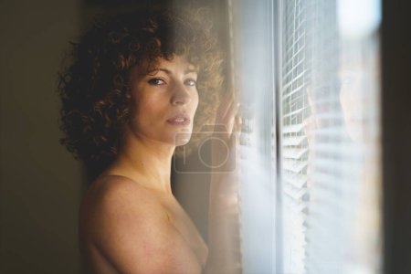 Foto de Sensual joven hembra desnuda con el pelo rizado de pie cerca de la ventana y mirando a la cámara en la sala de luz - Imagen libre de derechos