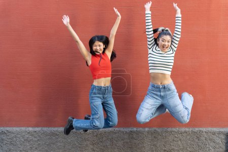 Foto de Cuerpo completo de amigas étnicas felices que usan jeans saltando con entusiasmo con los brazos levantados contra la pared roja mirando a la cámara - Imagen libre de derechos