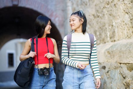 Téléchargez les photos : Heureux jeunes touristes asiatiques féminines en vêtements décontractés avec des sacs et un appareil photo se regardant tout en se promenant ensemble près d'un mur rugueux - en image libre de droit
