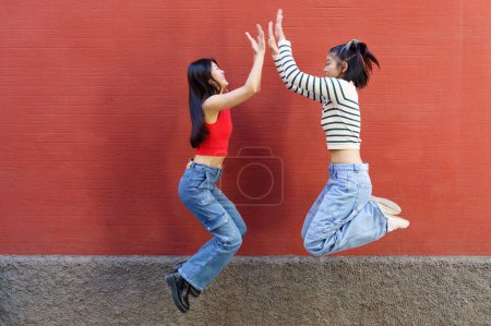 Téléchargez les photos : Plein corps vue latérale de joyeux jeunes asiatiques amies sautant vers le haut et donnant haute cinq à l'autre contre le mur rouge - en image libre de droit