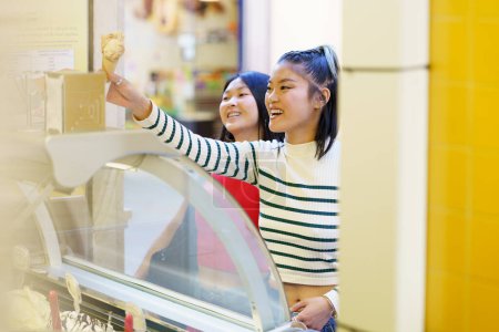 Photo for Positive young Asian girlfriends smiling happily while purchasing together ice cream in shop - Royalty Free Image