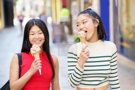 Photo for Happy Asian female friends eating delicious ice cream cone while walking together and enjoying time in city street - Royalty Free Image