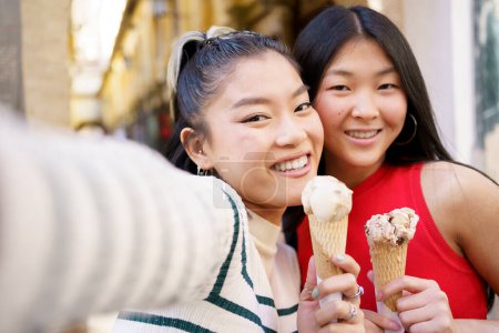 Photo for Positive Asian female friends taking self portrait while eating delicious ice cream cone against blurred background - Royalty Free Image