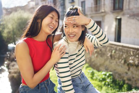Photo for Happy young Asian woman sitting with hand on shoulder of cheerful girlfriend while spending time together in Granada - Royalty Free Image