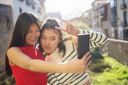 Photo for Happy Asian women in casual clothes showing rock gesture and using smartphone while shooting selfie together in Granada - Royalty Free Image