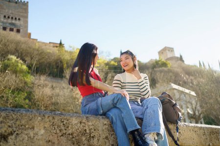 Photo for Happy young Asian female friends, in casual outfits smiling and looking at each other while talking on stone bench against ancient ruins of Paseo de los Tristes in Granada - Royalty Free Image