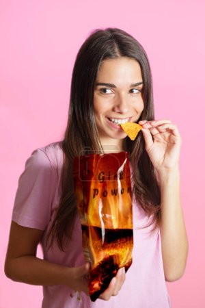 Photo for Positive young female in t shirt standing against pink background and looking away with biting delicious crunchy chips from pack with Girl Power words - Royalty Free Image