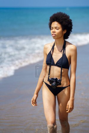 Foto de Grave fotógrafa afroamericana con peinado afro en traje de baño con cámara fotográfica caminando en la orilla cerca del mar en el día de verano - Imagen libre de derechos