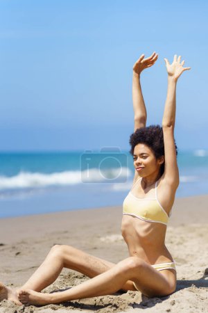 Photo for Content young fit African American female tourist with curly dark hair in stylish bikini stretching arms, while sitting on sandy beach against cloudless blue sky during summer vacation - Royalty Free Image