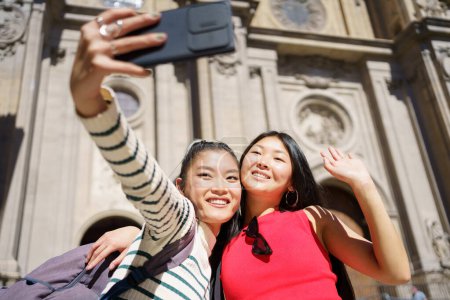 Téléchargez les photos : Heureux jeunes voyageuses asiatiques, en vêtements décontractés avec des sacs à dos souriants et câlins tout en agitant les mains et en prenant selfie sur smartphone pendant les vacances d'été en Espagne - en image libre de droit