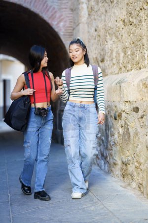 Foto de Cuerpo completo de jóvenes turistas asiáticas en ropa casual con bolsas y cámara fotográfica paseando contra la pared de piedra mientras hablan entre sí - Imagen libre de derechos