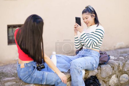 Téléchargez les photos : Jeune femme ethnique positive en pull rayé et en jeans, assise sur la frontière de pierre contre une petite amie aux longs cheveux noirs et prenant des photos sur smartphone pendant le voyage - en image libre de droit