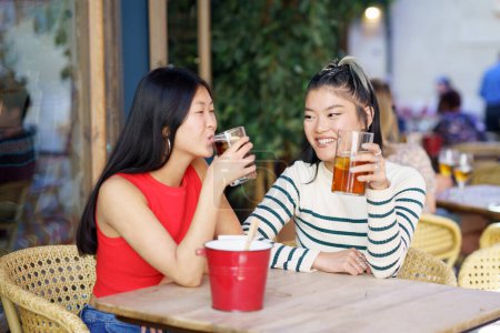 Foto de Alegre joven Asiática amigas en ropa casual sentado en la mesa de madera y charlando mientras bebe refrescante té frío durante la reunión en la cafetería - Imagen libre de derechos