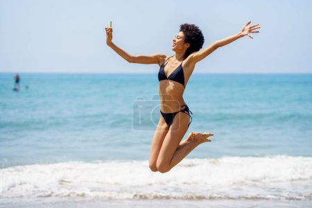 Photo for Full body side view of cheerful African American female traveler in bikini taking self portrait on smartphone and jumping on coast near waving sea - Royalty Free Image
