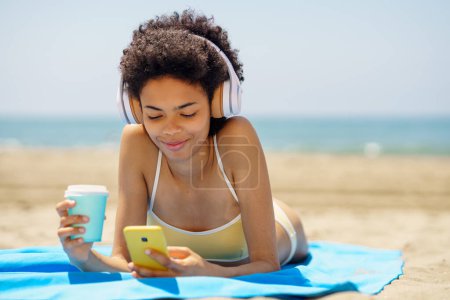 Photo for Content young ethnic female traveler with dark Afro hair, in stylish bikini and headphones, drinking takeaway coffee and messaging on smartphone while listening to music and sunbathing on sandy beach - Royalty Free Image