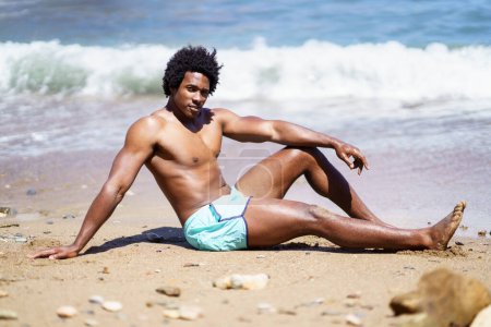 Téléchargez les photos : Vue latérale complète du corps d'un homme afro-américain se relaxant sur une plage de sable avec le bras sur le genou tout en regardant la caméra, et s'appuyant sur la main près de la mer mousseuse ondulante - en image libre de droit