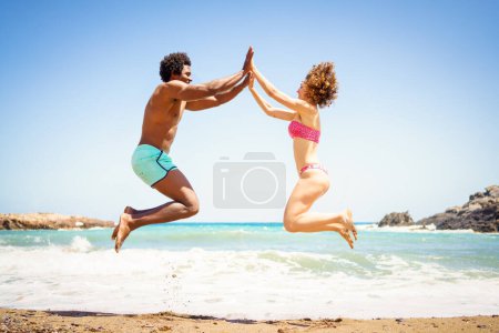 Téléchargez les photos : Pleine longueur vue latérale de l'homme et de la femme afro-américains joyeux en bikini sautant sur le bord de mer sablonneux et se tenant la main tout en se regardant - en image libre de droit
