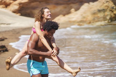 Foto de Alegre joven afroamericano hombre dando paseo a cuestas a la mujer de pelo rizado en bikini divertirse en la playa de arena cerca de mar ondulante - Imagen libre de derechos