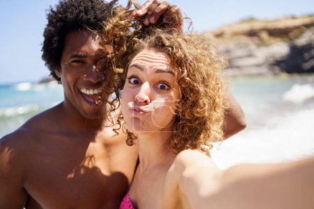 Photo for Cheerful young woman with puffed out cheeks taking selfie with black boyfriend touching curly hair of girlfriend while having fun on beach - Royalty Free Image