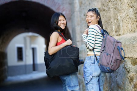 Photo for Cheerful Asian women tourists with belongings looking over shoulder at camera with backpack while standing near stone wall of building - Royalty Free Image