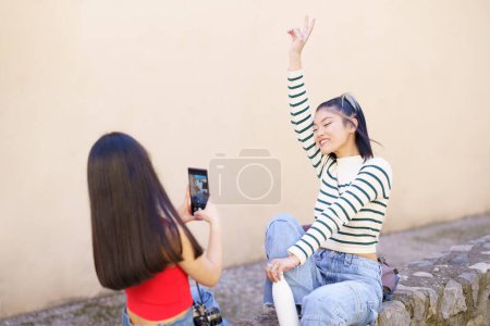 Photo for Happy young Asian women tourists taking picture on smartphone wile sitting on stone border on town street during vacation together - Royalty Free Image