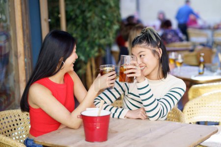 Foto de Positivas jóvenes amigas asiáticas en ropa casual sentadas juntas en la mesa de madera, y tintineando vasos de bebidas frescas mientras se miran durante la reunión en la cafetería - Imagen libre de derechos