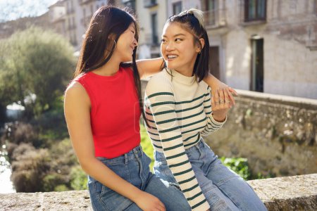 Photo for Positive Asian women smiling happily and looking each other while sitting on stone border of city in Granada - Royalty Free Image