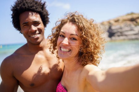 Foto de Alegre novia multirracial y novio en traje de baño sonriendo mientras toma selfie en día soleado cerca del mar durante las vacaciones de verano - Imagen libre de derechos