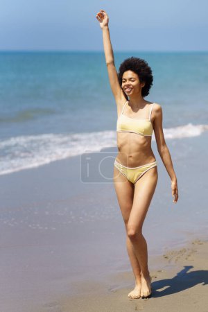 Photo for Full body of cheerful young African American female, traveler with dark curly hair in trendy bikini, smiling and looking away while walking on sandy beach with raised arm in sunlight - Royalty Free Image