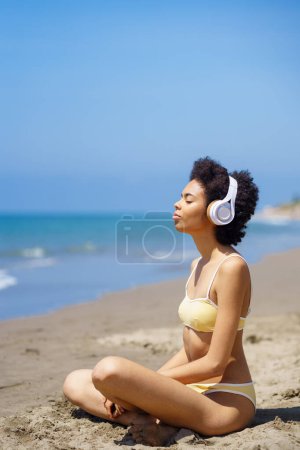 Photo for Side view of young fit ethnic female tourist with Afro hair in swimwear listening to music in headphones, while meditating on sandy beach with closed eyes in Lotus pose - Royalty Free Image