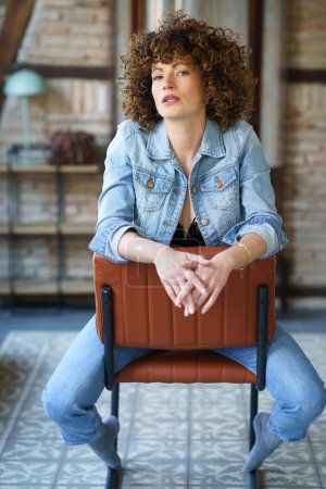 Photo for Serious young woman wearing denim jacket sitting on leather chair and touching hair while looking at camera against blurred brick wall - Royalty Free Image