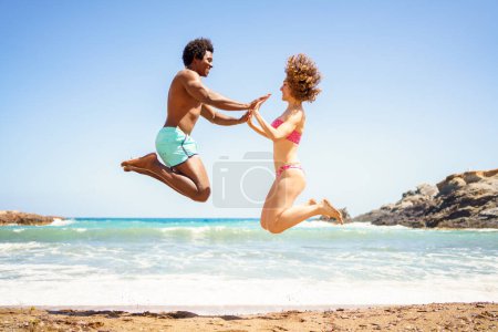 Téléchargez les photos : Vue latérale complète du corps de joyeux petit ami afro-américain et petite amie aux cheveux bouclés, en maillot de bain sautant sur la plage de sable et se tenant les mains près de la mer tout en se regardant - en image libre de droit