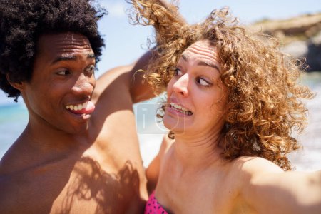Téléchargez les photos : Homme noir joyeux avec des cheveux afro et la langue toucher les cheveux bouclés de petite amie faire grimace drôle sur une journée ensoleillée près de la mer - en image libre de droit