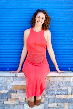 Téléchargez les photos : Corps complet de jeune femme relaxante en robe de soleil rouge assis avec les mains sur le grès et souriant tout en regardant la caméra contre les volets bleus en plein jour - en image libre de droit